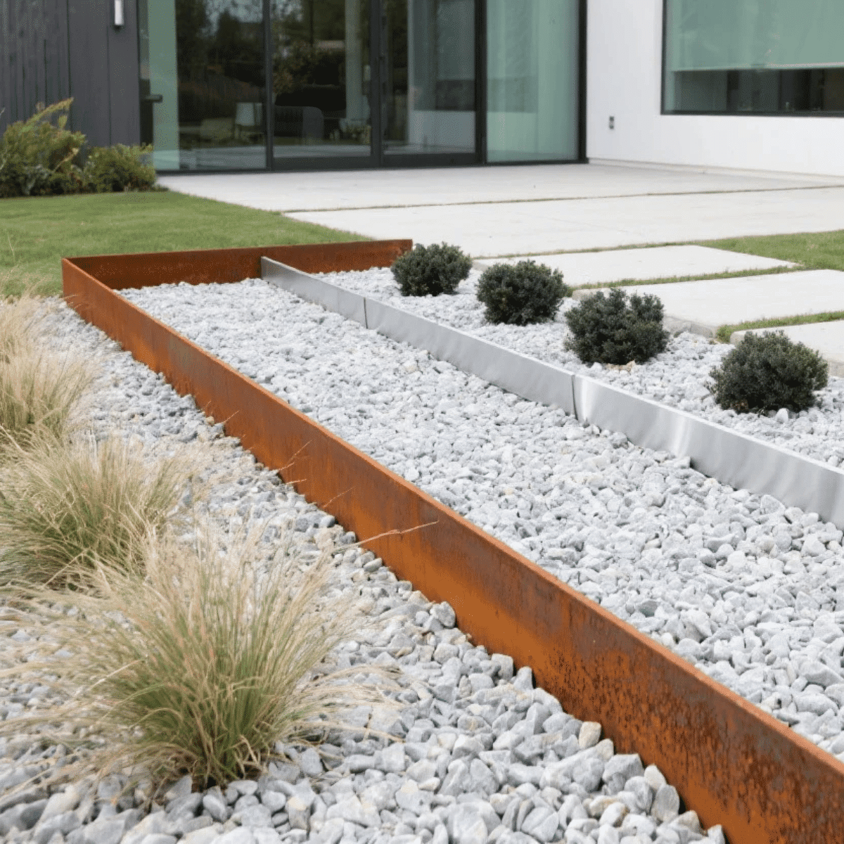 Moderne Gartengestaltung mit rostfarbener Stahl-Beeteinfassung, hellem Kies, Ziergräsern und kugelförmig geschnittenen Sträuchern vor einem Haus.